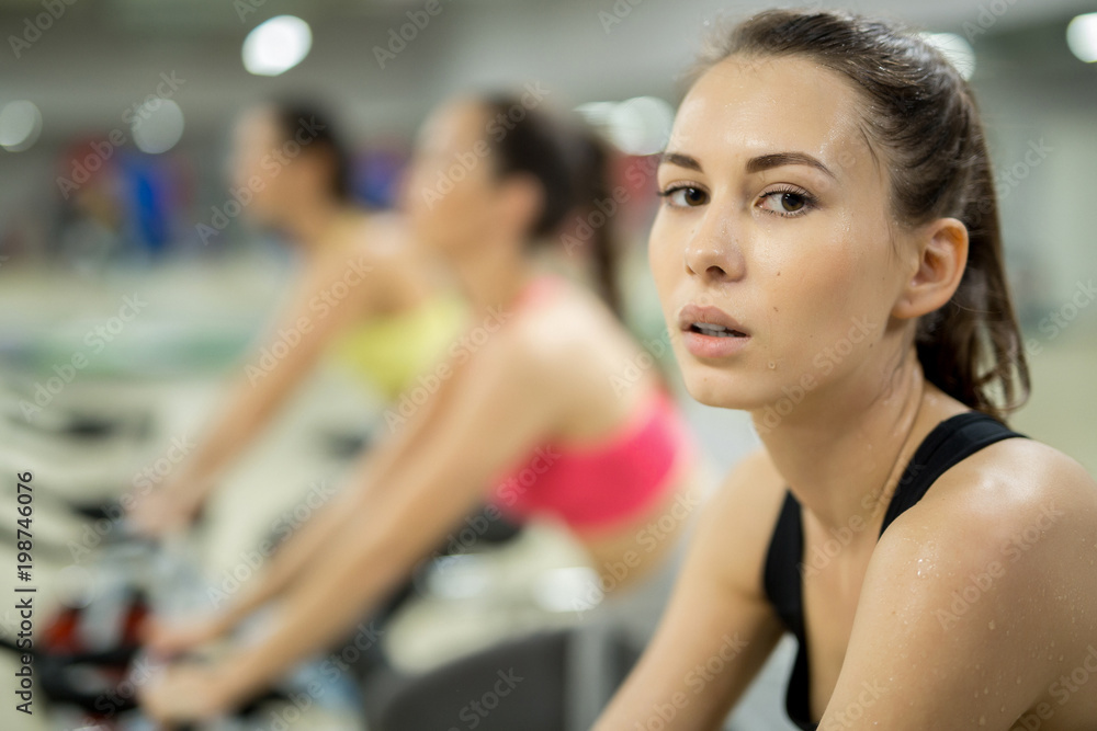 Wet young woman training hard on sports bike in gym on background of her friends