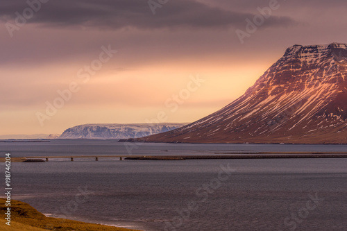 view of bridge along cliff and ocean with traffic and big mountain in background. Sea Cliff Bridge view on dramatic sky on the way to Grundarfjordur in Iceland.