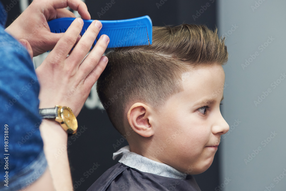 Fototapeta premium Barber is using styling gel and hairbrush to make a styling to a Caucasian boy.