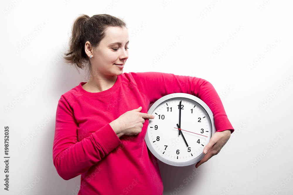 brunette girl is holding clock and points to hands of clock on white background. concept of time. Watch, stop and control time. Young woman holds round clock in hands. brunette girl is holding clock and points to hands of clock on white background. concept of time. Watch, stop and control time. Young woman holds round clock in hands.