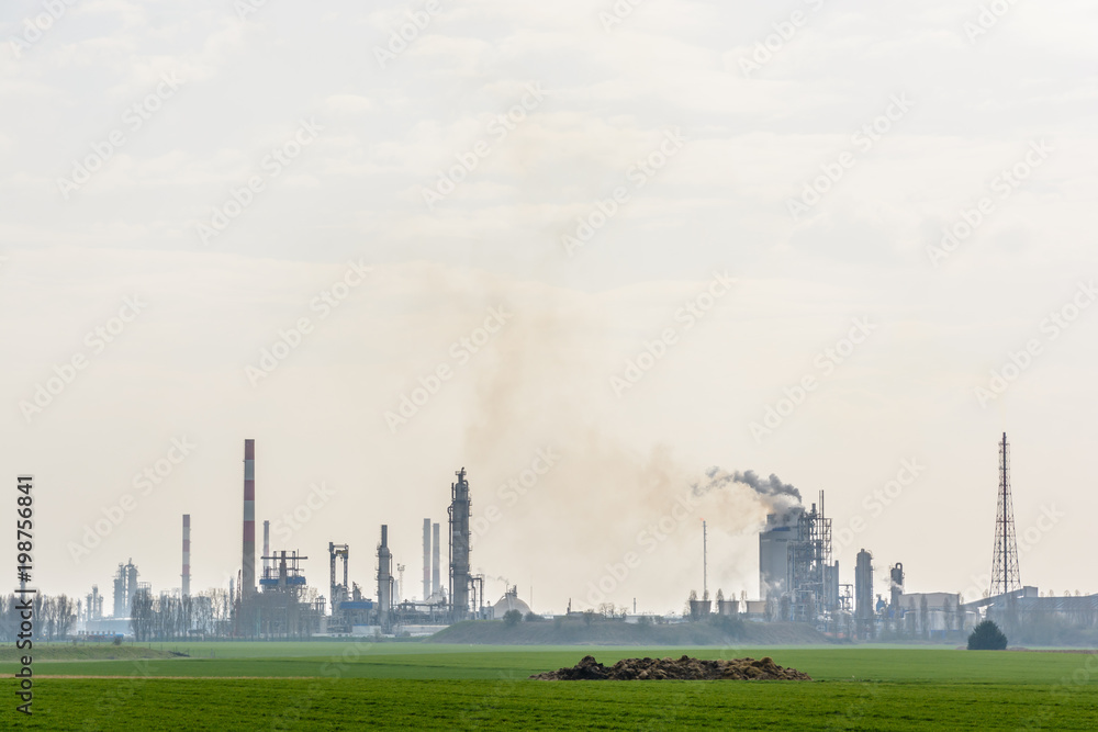 View of an oil refinery with smoking chimneys releasing toxic gases in the air, surrounded by crop fields in the french countryside under a pale sunlight. View of an oil refinery with smoking chimneys releasing toxic gases in the air, surrounded by crop fields in the french countryside under a pale sunlight.