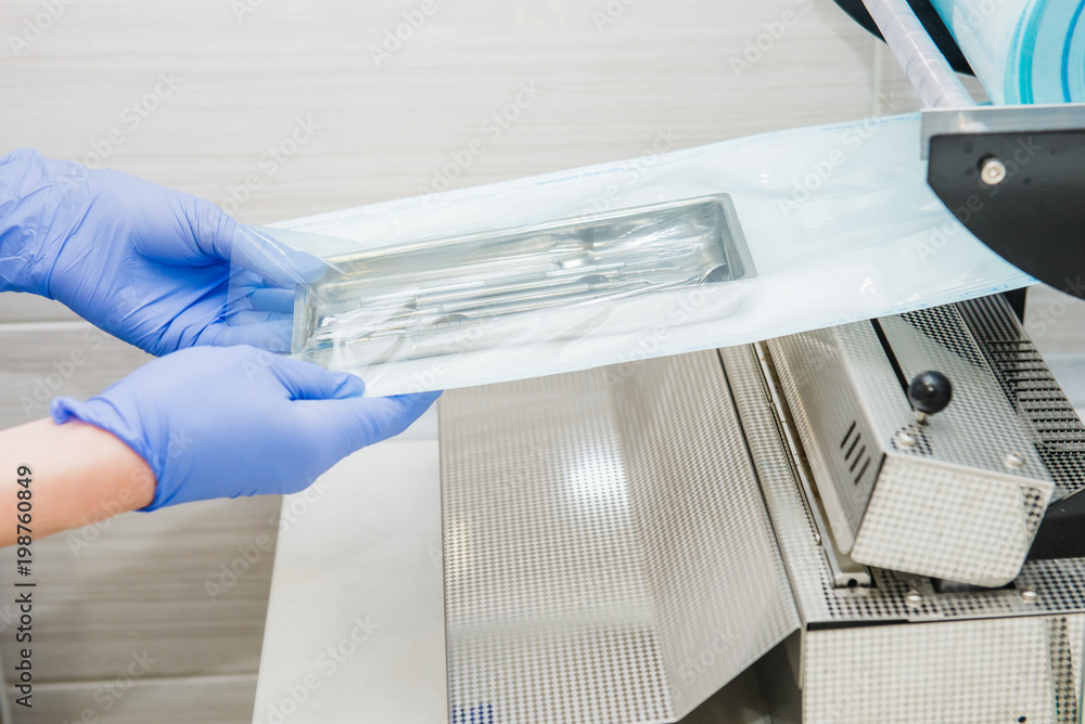 Close up dentist's assistant hands in gloves packing dental instrument set for autoclaving in a plastic bag using vacuum packing machine. Sterility and safety care concept. Selective focus. Close up dentist's assistant hands in gloves packing dental instrument set for autoclaving in a plastic bag using vacuum packing machine. Sterility and safety care concept. Selective focus.