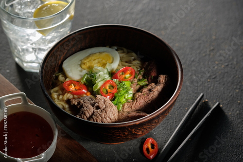 Asian ramen soup with beef, egg, chives in bowl on grey background.