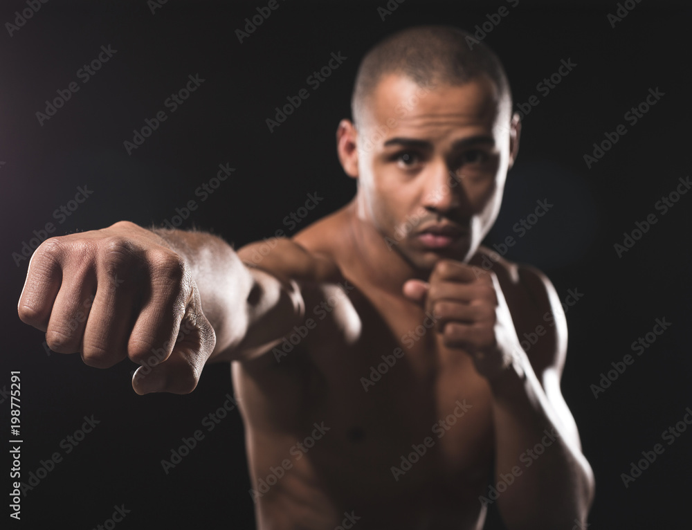Waist up portrait of young naked african guy looking pending at camera while beating by right fist in front of him. Focus on combat hand. Isolated on black background