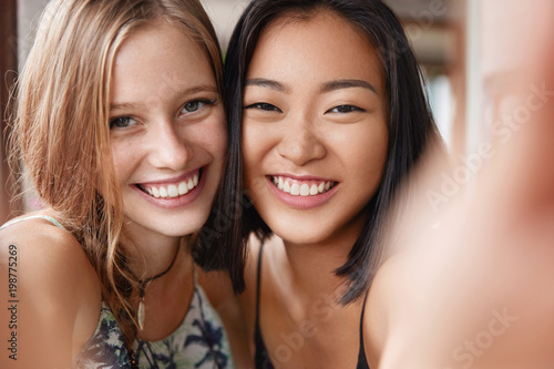 Happy European female embraces best Asian friend, make selfie, stand close to each other. Positive young women bloggers shoot video how they rest during free time. Interracial friendship concept