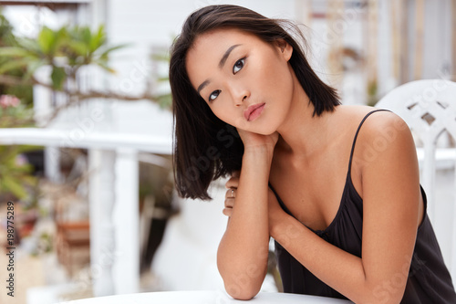 Adorable serious Asian female has serious expression, poses at cafe alone, feels bored in unknown country, dressed casually, has good recreation. Relaxed mixed race woman enjoys leisure time