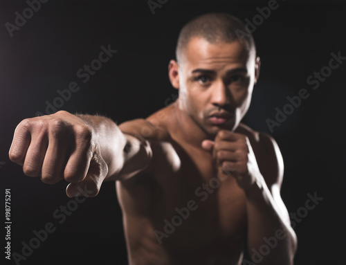 Waist up portrait of young naked african guy looking pending at camera while beating by right fist in front of him. Focus on combat hand. Isolated on black background