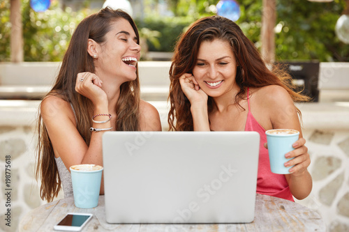 Joyful women have fun together, laugh while watch comedy online on laptop computer and recreat in outdoor cafe, drink hot coffee or espresso, enjoy free time and vacations. People, technology photo