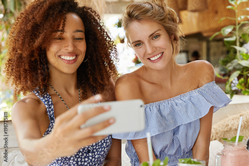 Mixed race friendly females have positive expressions, pose for selfie in modern smart phone, have satisfied looks, recreat in coffee shop together. African American woman takes photo on cellular photo