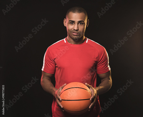 Wait up portrait of young African man in red t-shirt with basketball. He looking at camera and smiling barely noticeable. Isolated on black background