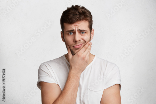 Photo of upset male with stubble, dressed in white t shirt, curves face, isolated over white background. Discontent young man wears casuual clothing, dislikes something. People and emotions concept