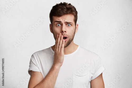 Tableau sur toile Photo of shocked handsome man with attractive look, stares at camera in bewilderment, hears bad news, poses in studio against white background