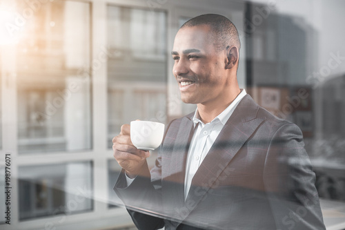 Waist up portrait of handsome businessman looking into the distance with happy smile while savour cup of tea. Man is on the other side window of cabinet