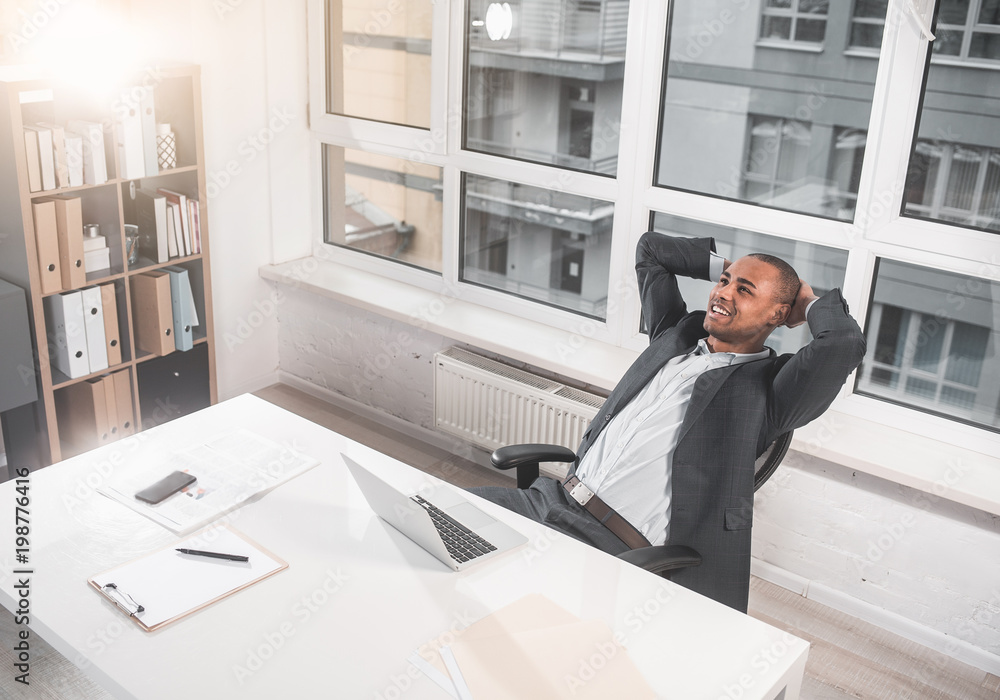 Top view of young african guy is sitting at office in chair and reclining on the back. He smiling while with relaxation looking at ceiling. Copy space in left side Top view of young african guy is sitting at office in chair and reclining on the back. He smiling while with relaxation looking at ceiling. Copy space in left side