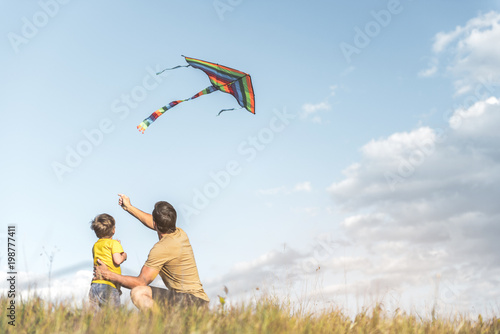 Parent and little boy are sitting with their backs to camera on field and flying kite. Copy space in right side