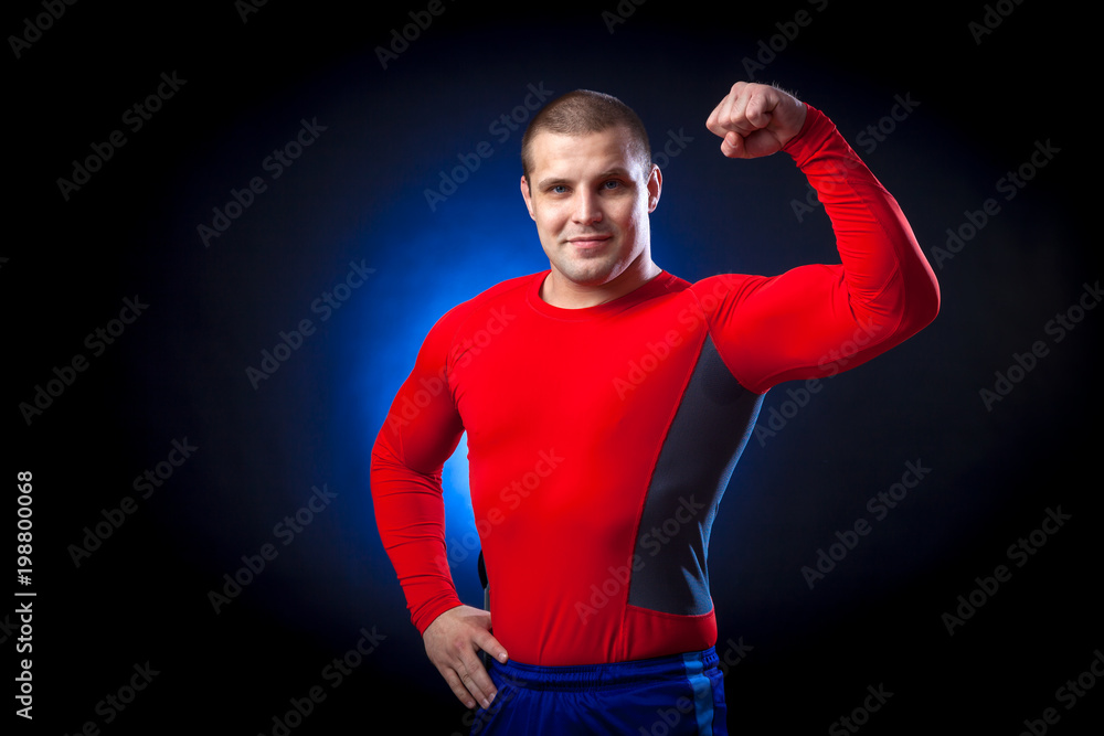 A strong dark-haired sportman  in a red sports wear  rush guard shows  biceps against a blue a lights on a black isolated  background