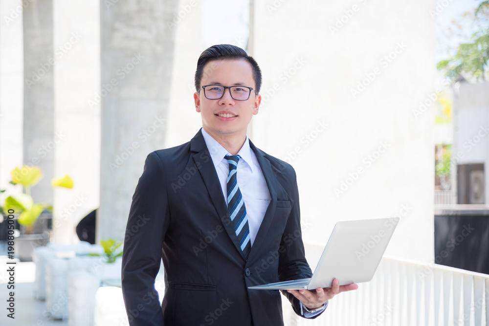 Young asian businessman in suit holding laptop computer and smiles with modern building on background Young asian businessman in suit holding laptop computer and smiles with modern building on background