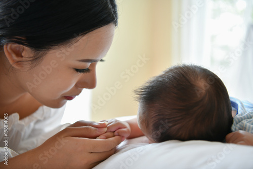 Newborn Concept. Mother and child on a white bed. Mom and baby boy playing in bedroom. Parent and little kid relaxing at home. Family having fun together. Newborn baby is fussing and crying.