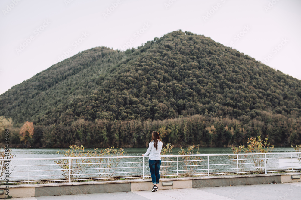 Young girl stands beside a beautiful lake. In the back there is a green hill