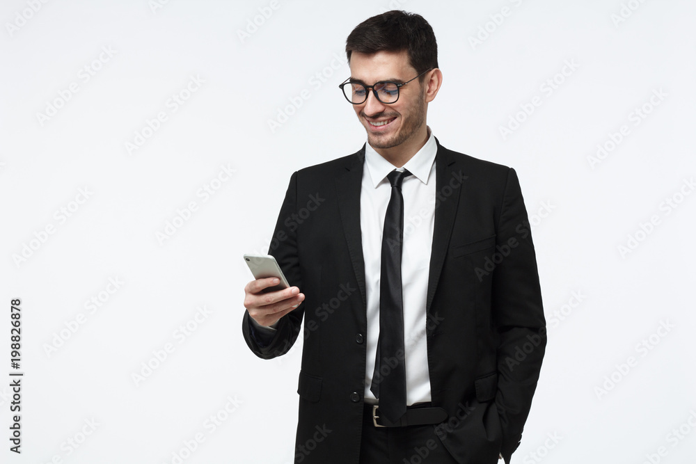 Positive smiley pleased satisfied salesman looking at smartphone screen, checking currency rates, isolated on grey background Positive smiley pleased satisfied salesman looking at smartphone screen, checking currency rates, isolated on grey background