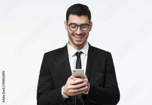 Young business man laughing, looking at mobile phone display, holding his smartphone with both hands, isolated on grey background