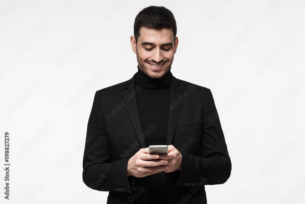 Young business man standing isolated on grey background looking attentively at screen of cellphone, browsing web pages and smiling nicely while chatting Young business man standing isolated on grey background looking attentively at screen of cellphone, browsing web pages and smiling nicely while chatting