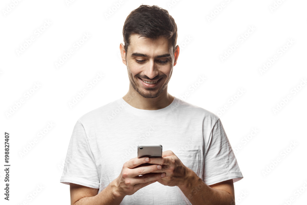 Young man in blank tshirt standing isolated on white background looking attentively at screen of cellphone, browsing web pages and smiling nicely while chatting with friends Young man in blank tshirt standing isolated on white background looking attentively at screen of cellphone, browsing web pages and smiling nicely while chatting with friends