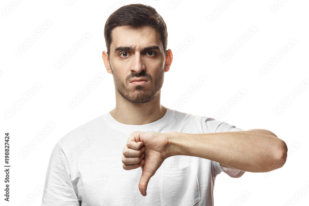 Disgruntled grumpy dissatisfied annoyed unhappy young man showing thumb down as dislike gesture isolated on white background Disgruntled grumpy dissatisfied annoyed unhappy young man showing thumb down as dislike gesture isolated on white background