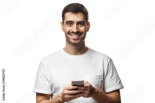 Portrait of handsome young man in white t-shirt, holding smartphone, looking at camera and smiling