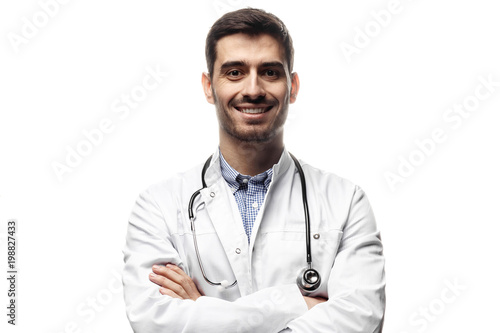 Closeup shot of young caucasian man doctor standing with crossed arms in white uniform against white background looking perfectly confident © Damir Khabirov