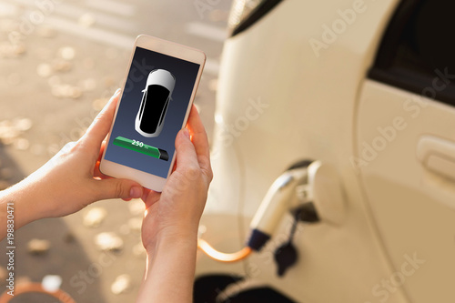 Hands with phone on a background of charging electric car. On a device screen indicator of power reserve. 