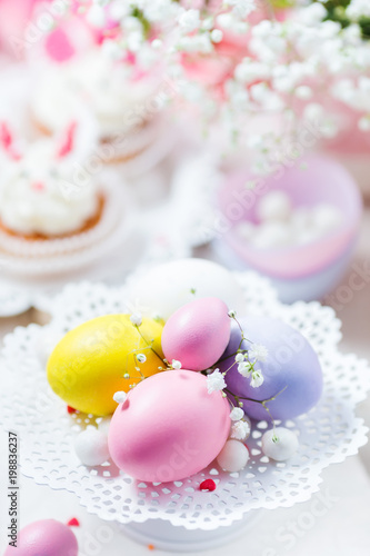 Easter eggs in pastel color on a white cake stand, bunny cupcakes and flowers. Festive light background