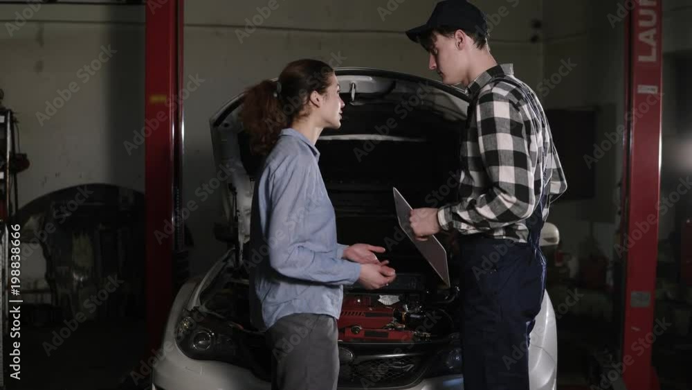 Cute car or automotive technician wearing work robe and a cap is meeting with his female clients and hears out complaints about her vehicle. Cute car or automotive technician wearing work robe and a cap is meeting with his female clients and hears out complaints about her vehicle.