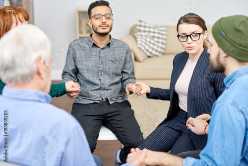 Fototapeta Naklejka Na Ścianę i Meble -  Multi-ethnic group of patients suffering from phobias sitting in circle and holding hands while highly professional psychologist conducting session, interior of cozy office on background