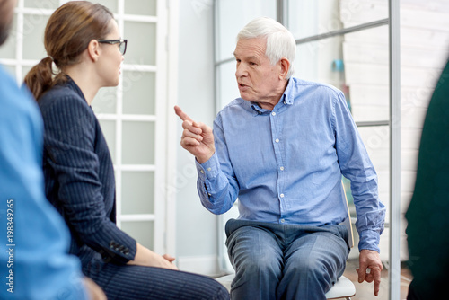 Portrait shot of senior patient communicating with highly professional psychologist while participating in group therapy session, interior of psychotherapy office on background