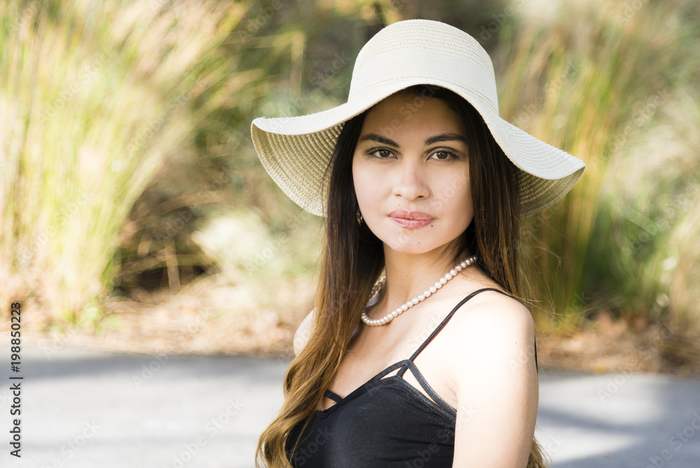 Outdoor portrait of a young woman in white hat. Attractive woman in park, blurred plant in background.