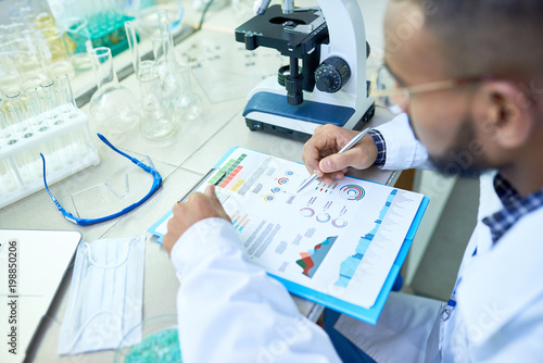 Close-up of busy concentrated Arabian male scientist viewing charts while holding medical research and sitting at table with glassware and equipment in modern laboratory