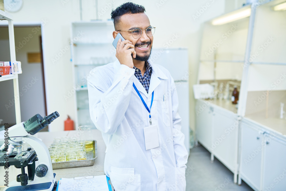 Cheerful excited handsome Arabian laboratory employee with hanging badge on neck communication with colleague on mobile phone and laughing Cheerful excited handsome Arabian laboratory employee with hanging badge on neck communication with colleague on mobile phone and laughing
