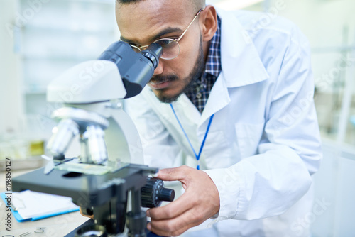 Serious concentrated young Arabian male scientist using fine focus knob of microscope to bring image into focus in laboratory
