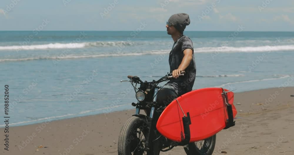 custom made wallpaper toronto digitalHandsome man biker surfer standing next to black motorcycle cafe racer with red surfboard shortboard on beach at sunny day and looking waves. 4k video shooting by handheld gimbal