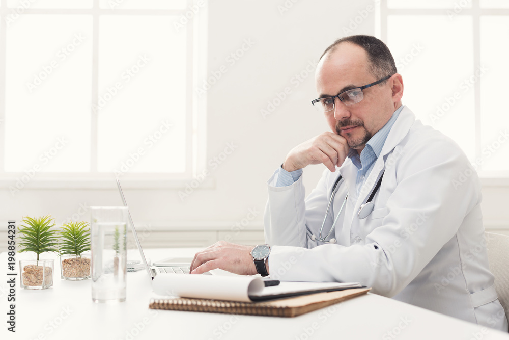 Serious doctor in glasses typing on laptop