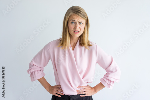 Portrait of young Caucasian businesswoman wearing pink blouse standing with hands on waist displeased at some rude behavior. Irritation, misunderstanding concept