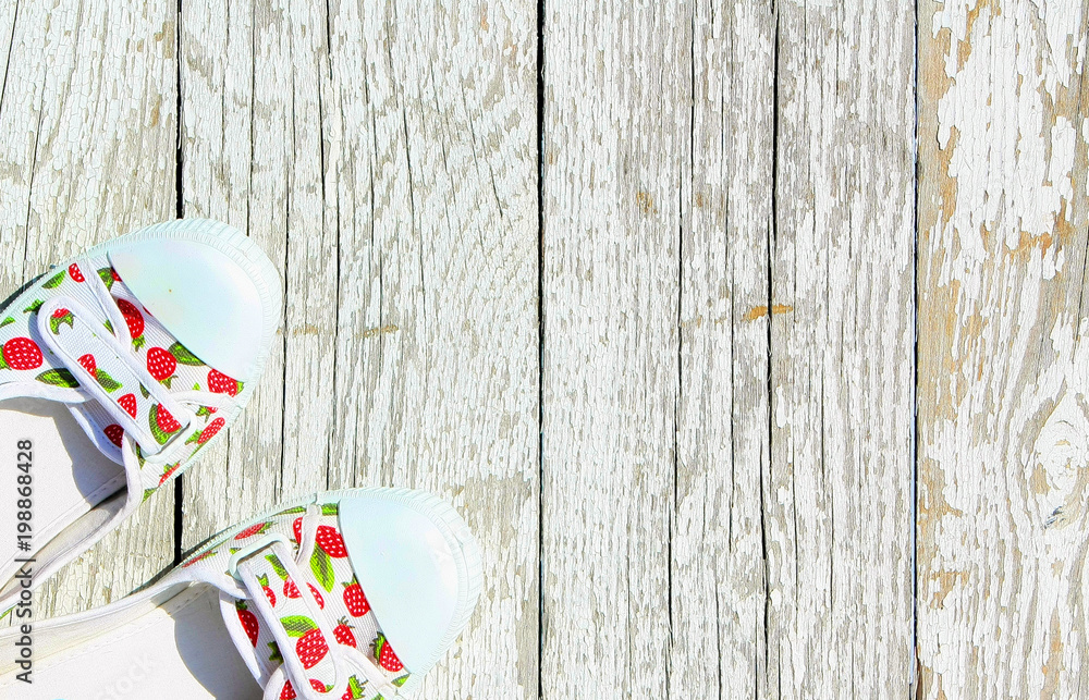 Obraz premium White with red berries sneakers on a white wooden background. Summer shoes top view. Colored slippers stand on old painted boards. Free space for text.