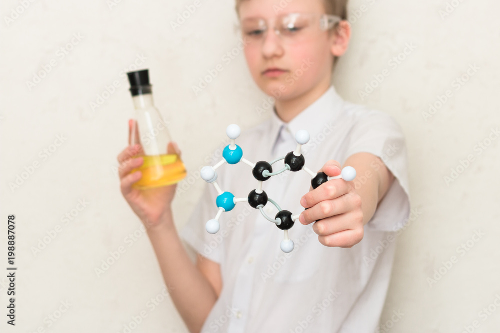 A teenager boy in a chemistry class holds a flask with chemicals and a plastic model of a molecule. Science. STEM education. School lab. A teenager boy in a chemistry class holds a flask with chemicals and a plastic model of a molecule. Science. STEM education. School lab.