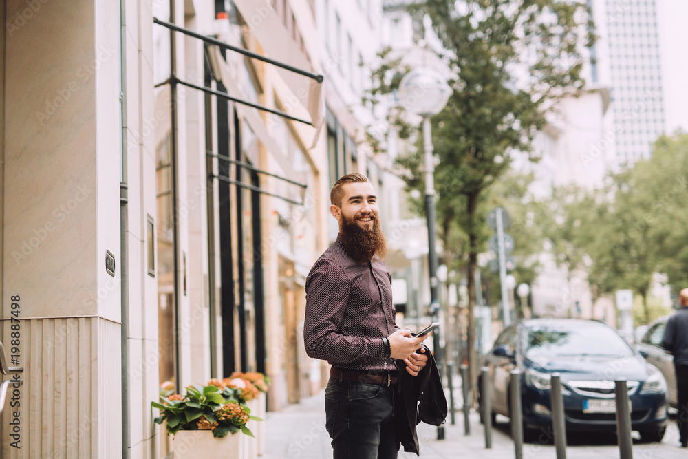 Young man with the beard uses a mobile phone in the city beside the office Young man with the beard uses a mobile phone in the city beside the office