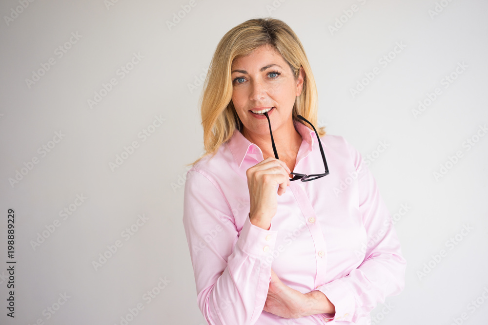 Closeup portrait of content middle-aged attractive fair-haired woman looking at camera and biting glasses temple. Smart middle-aged woman concept. Isolated front view on grey background. Closeup portrait of content middle-aged attractive fair-haired woman looking at camera and biting glasses temple. Smart middle-aged woman concept. Isolated front view on grey background.