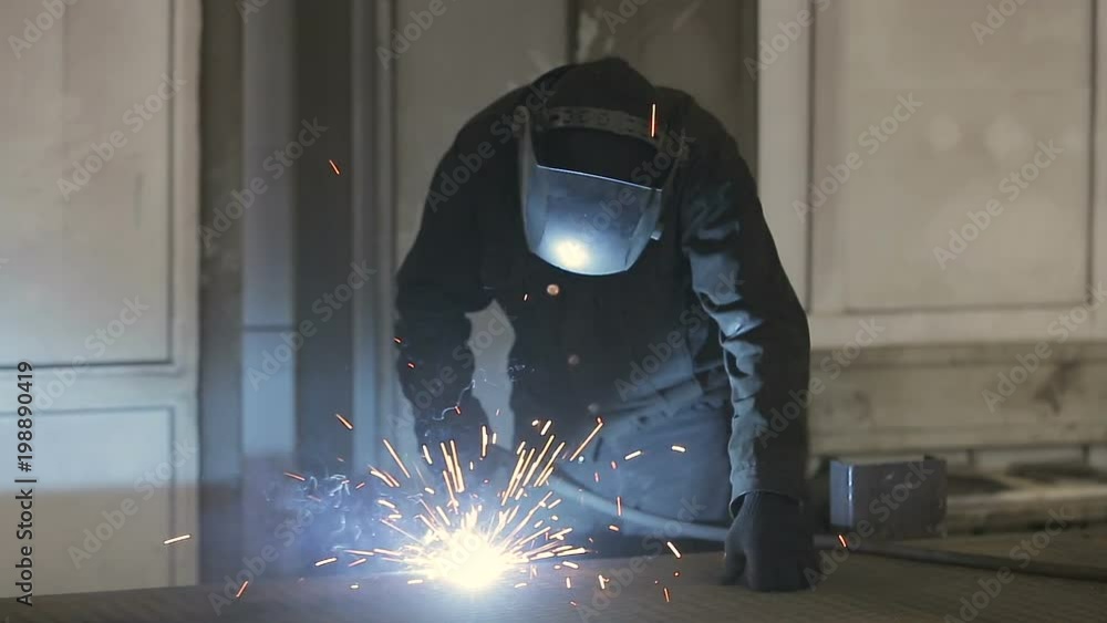 The working welder processes the metal, prepares the base. A man in a protective suit, sparks. The working welder processes the metal, prepares the base. A man in a protective suit, sparks.