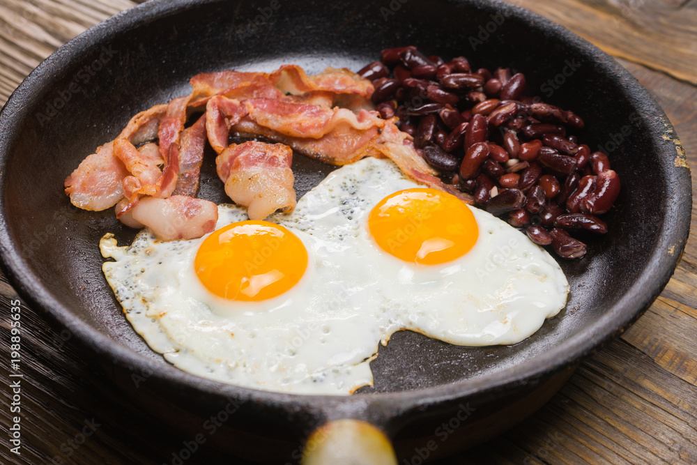 Bacon,Egg and bean. Salted egg and sprinkled with black pepper. English breakfast. Grilled bacon, two eggs and beans in pan on wooden table Bacon,Egg and bean. Salted egg and sprinkled with black pepper. English breakfast. Grilled bacon, two eggs and beans in pan on wooden table