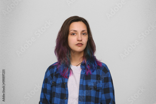 Indoor shot of young caucasian girl wears checkered blue shirt with dyed colorful hair looks directly into camera. Serious expression. Isolated over white wall. © timtimphoto