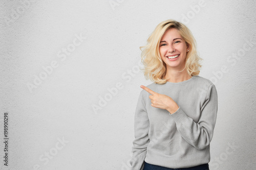 Happy excited female with blonde hair indicates with fore finger aside, demonstrates something at blank copy space for your advertisment or hearder. Positive expression. Isolated photo